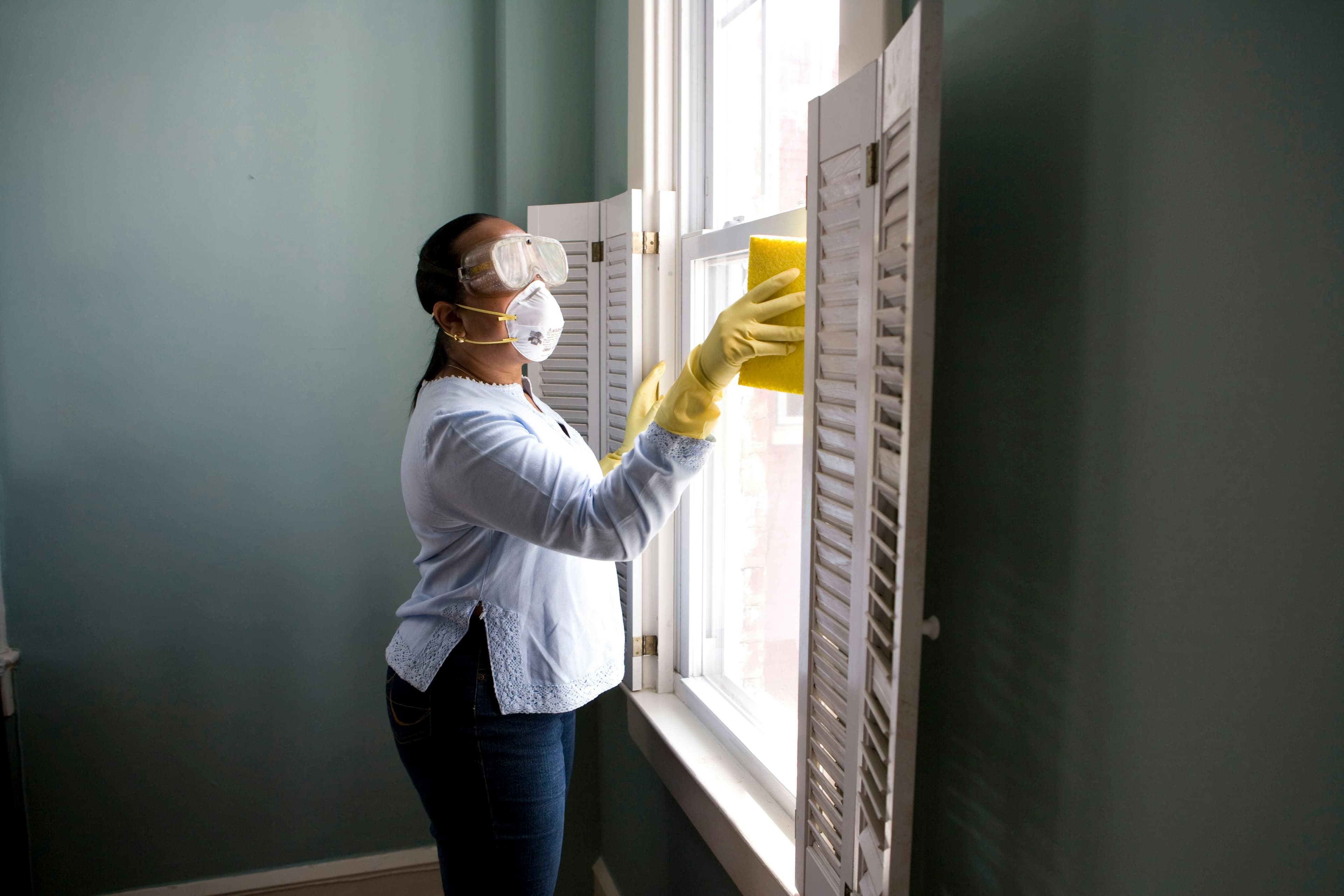 Cleaner wiping down interior window shutters in a bright room